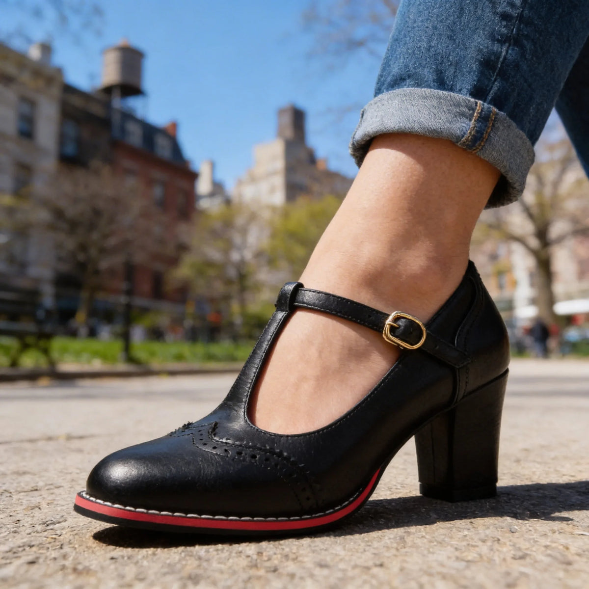Close-up of black T-strap heel with gold buckle, wingtip stitching and block heel on city street