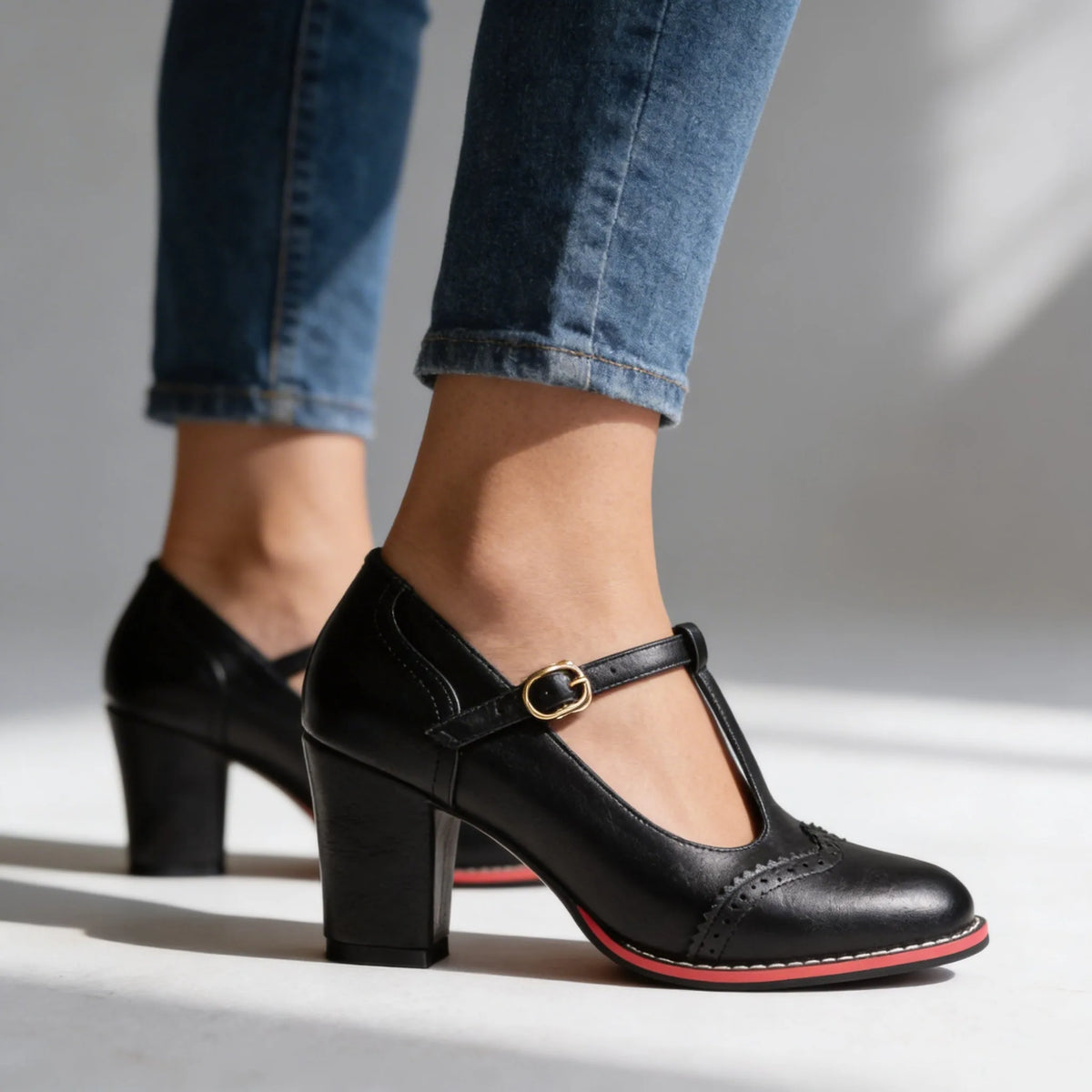 Black leather T-strap brogue pumps with red trim sole shown walking against a studio backdrop