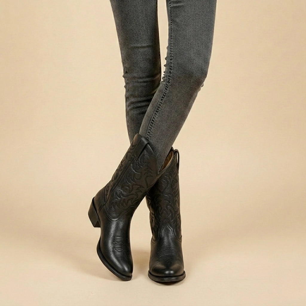 Front view of black leather pull-on western boots with scalloped shaft and tonal stitching, worn with grey skinny jeans