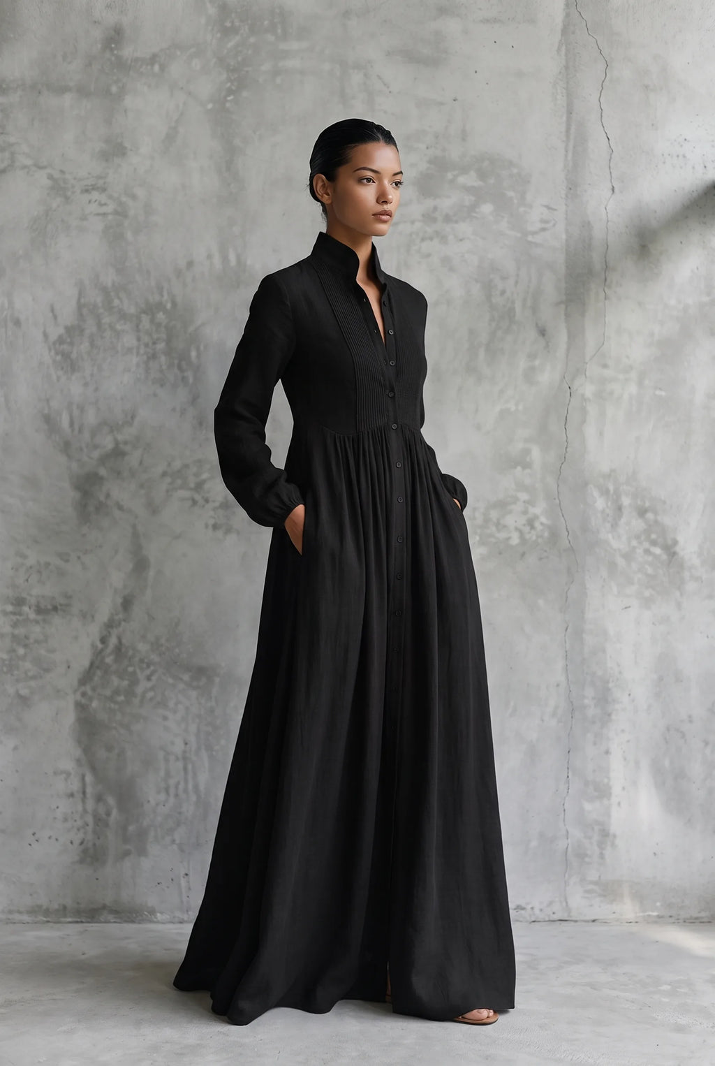 Three-quarter view of a model in a black cotton linen maxi dress, hands in pockets, showing the flowing A-line skirt.