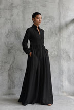 Three-quarter view of a model in a black cotton linen maxi dress, hands in pockets, showing the flowing A-line skirt.