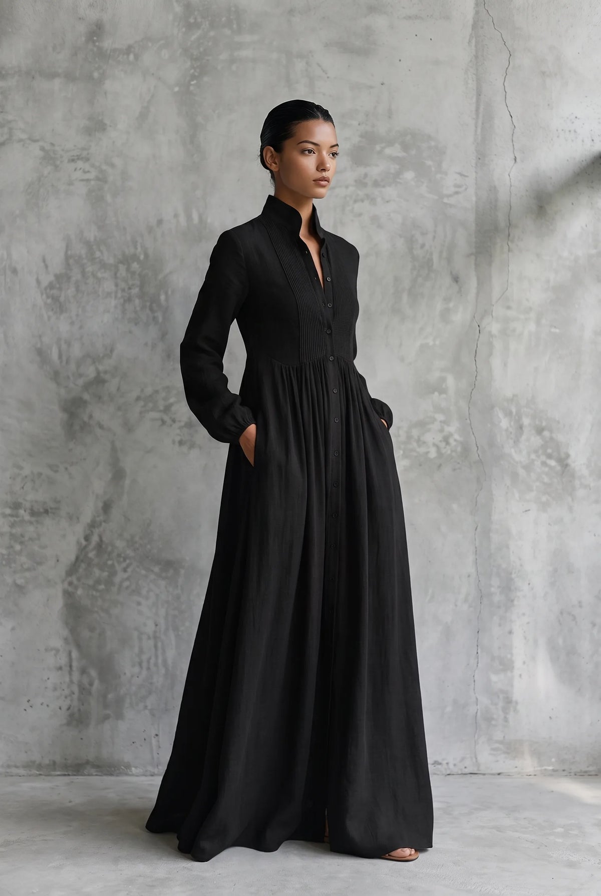 Three-quarter view of a model in a black cotton linen maxi dress, hands in pockets, showing the flowing A-line skirt.