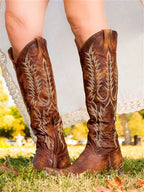 Back view of brown leather knee-high western boots with cream embroidery and low stacked heel, worn with a flowing dress