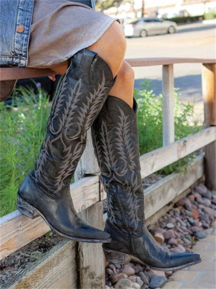 Side angle of black leather knee-high boots worn while seated on a wooden rail, highlighting pointed toe shape