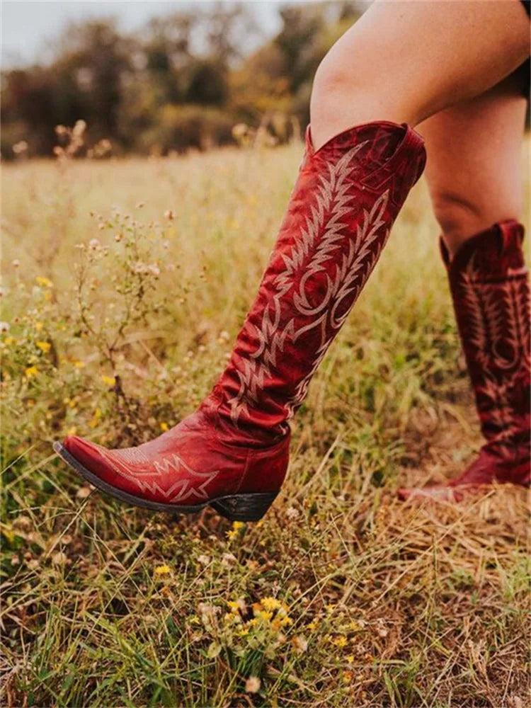 Walking shot of red leather knee-high western boots in a grassy field showing the embroidered shaft in motion