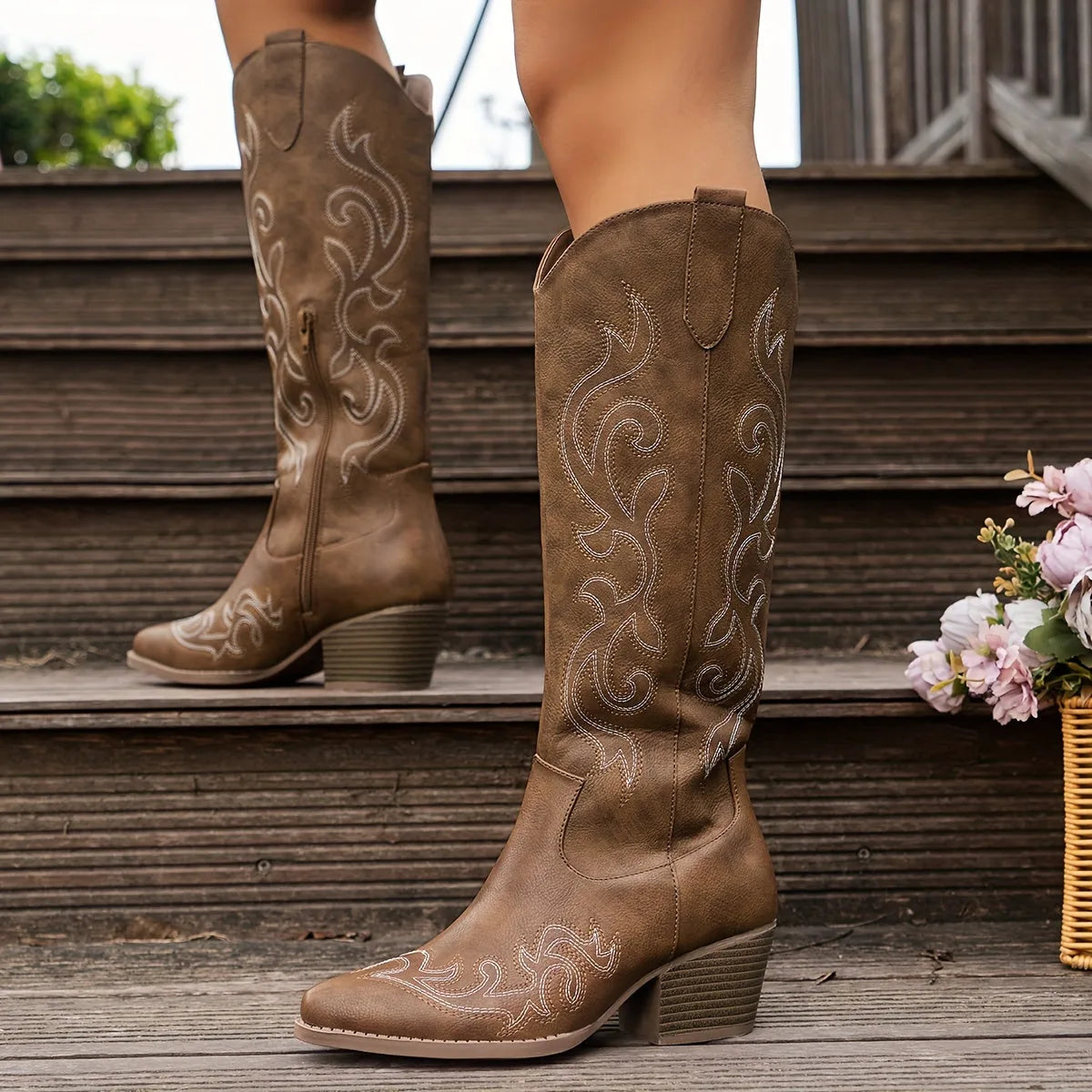 Side view of brown knee-high cowboy boots with cream stitching worn on a wooden staircase
