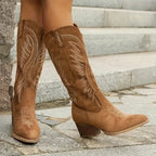 Side view of tan suede knee-high cowboy boots with embroidered stitching and a stacked block heel, worn on stone steps.