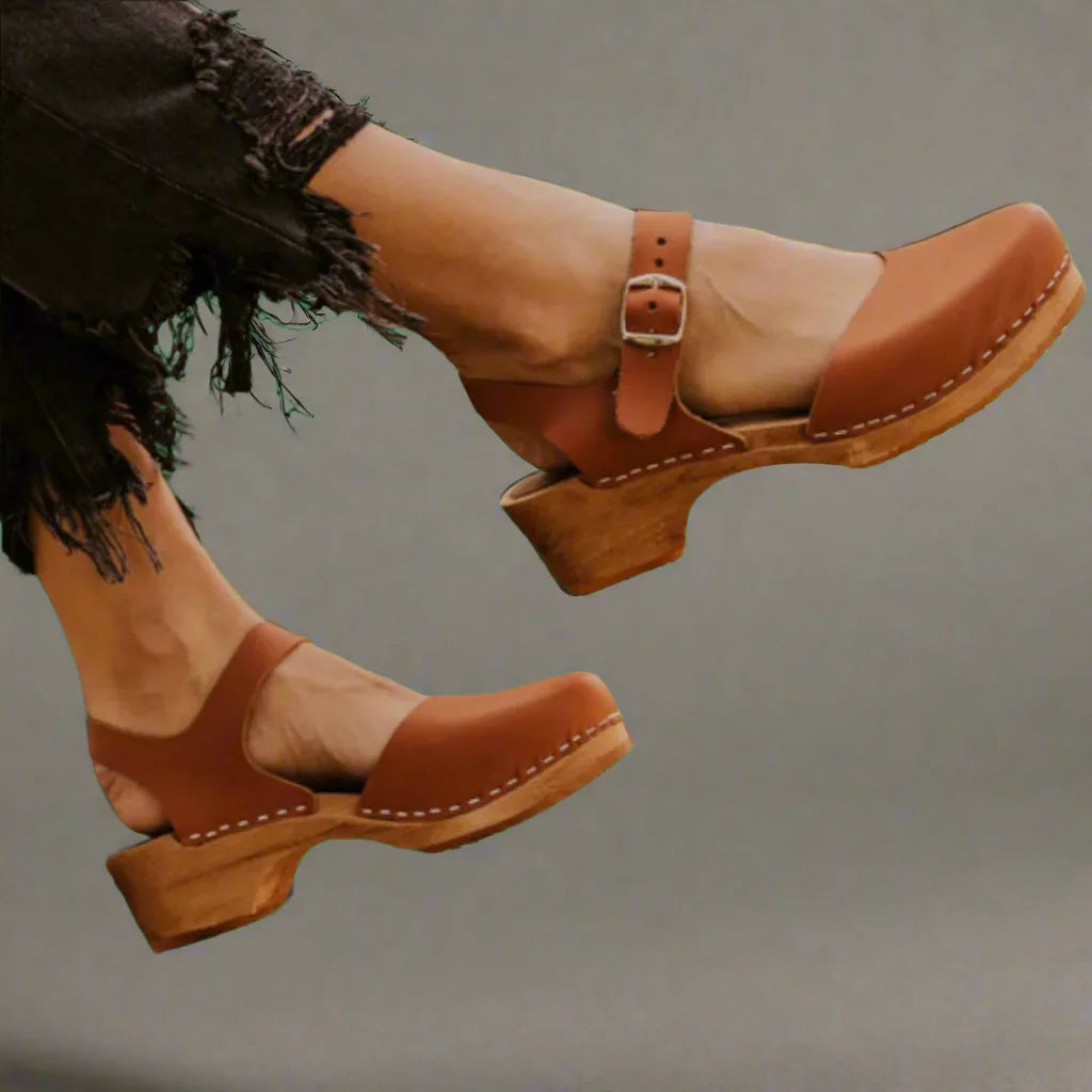 Side view of tan leather clog sandals with buckled strap, round toe and stacked wooden heel, worn on bare feet