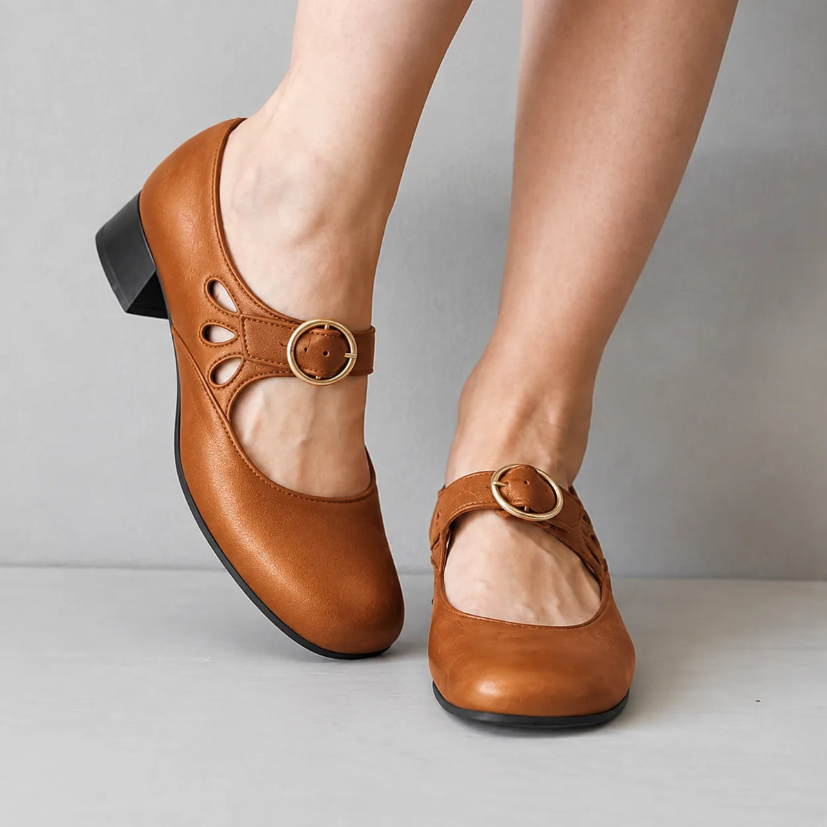 Tan leather mary jane flats with cutout detailing and a gold round buckle, paired on crossed legs