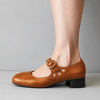 Close side profile of a tan leather mary jane flat showing the round gold buckle and low stacked heel