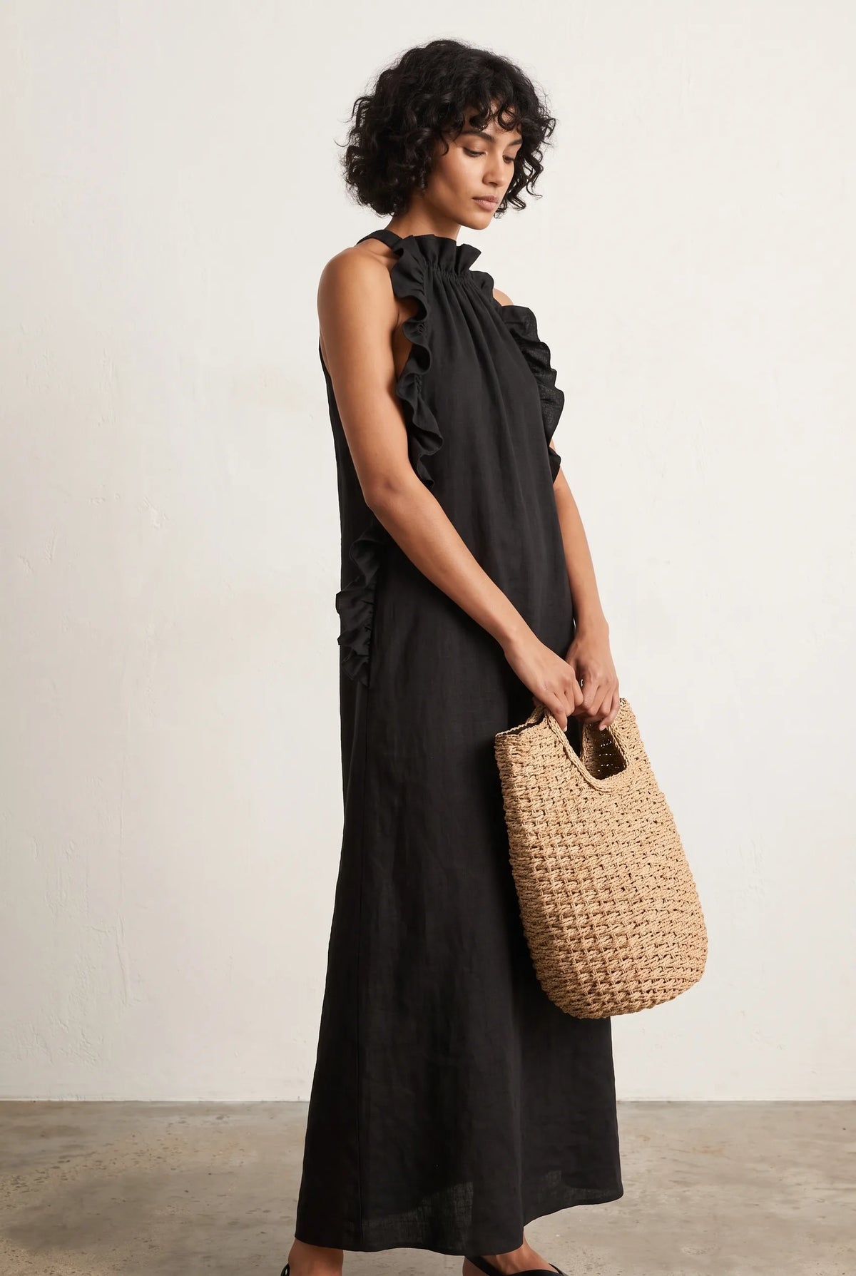 Front view of a model in a black linen halter maxi dress with ruffled neckline and ties, holding a woven straw bag