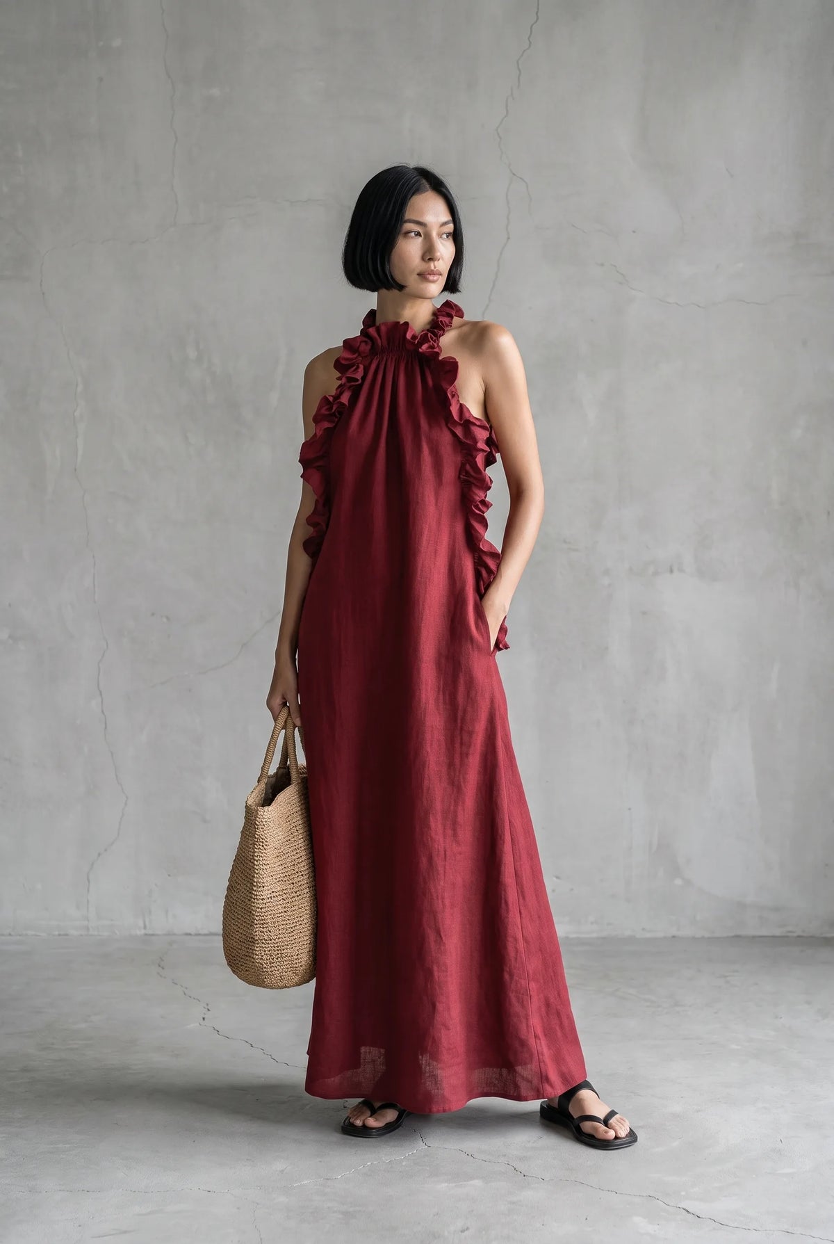 Front view of a model wearing a deep red linen halter maxi dress with ruffled trim down the bodice and a straw tote