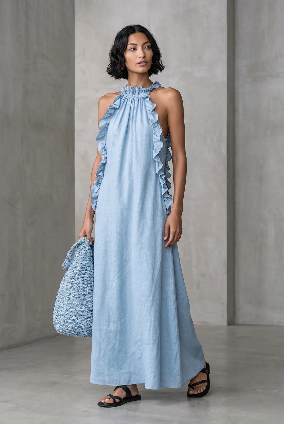 Model wearing a light blue linen-blend halter maxi dress with ruffled edges, paired with a matching woven handbag