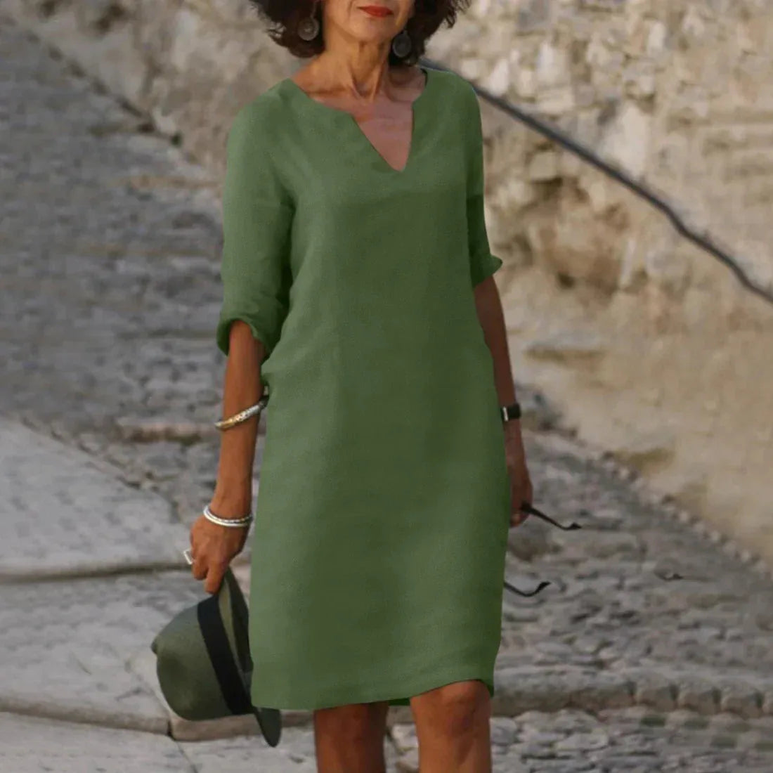Olive green linen-style dress with notched neckline, shown walking outdoors near a stone wall