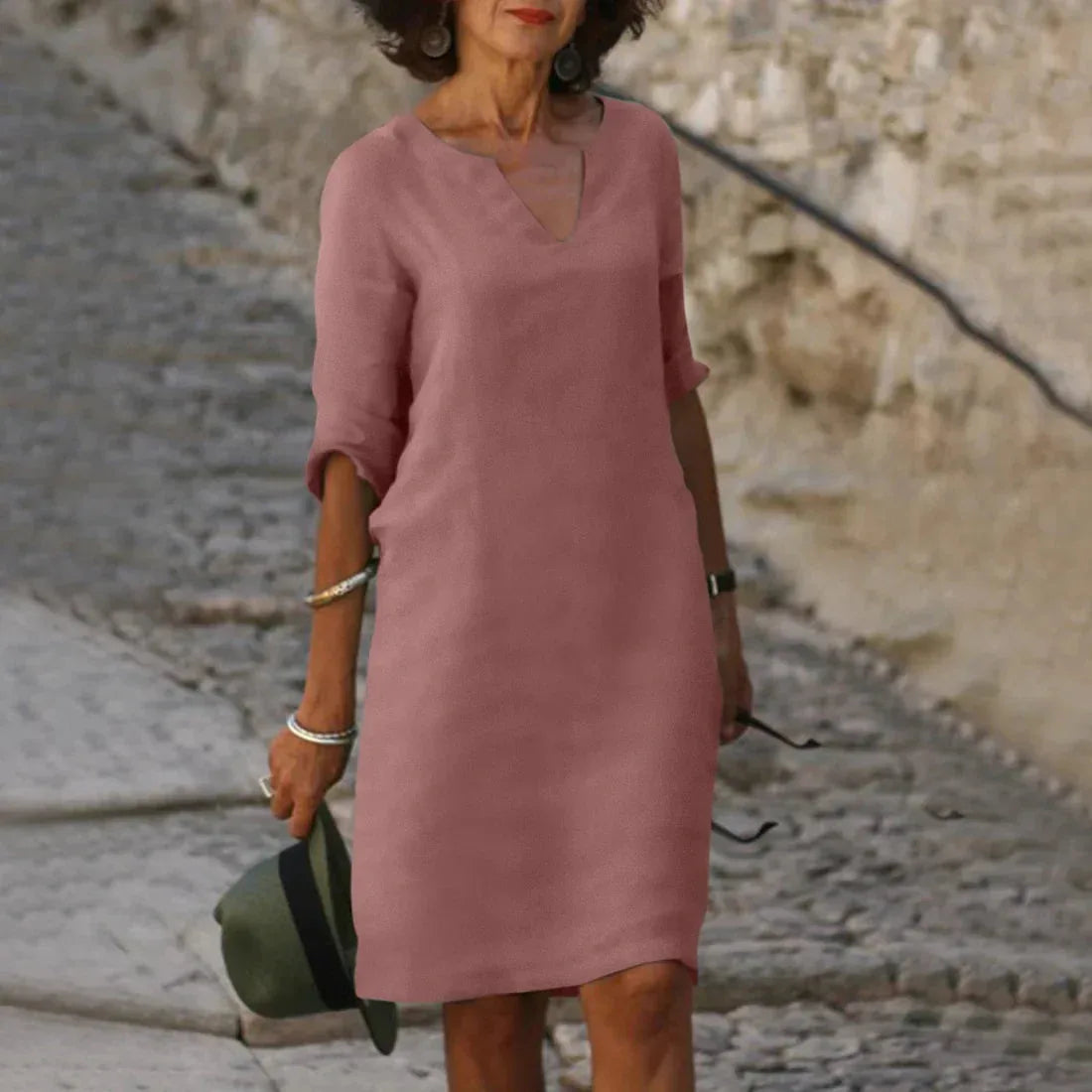 Dusty pink V-neck shift dress with three-quarter turned sleeves, worn against a sunlit stone backdrop
