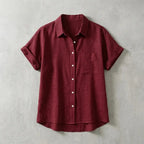 Folded burgundy red linen short-sleeve shirt with chest pocket and mother-of-pearl buttons, laid flat on concrete background
