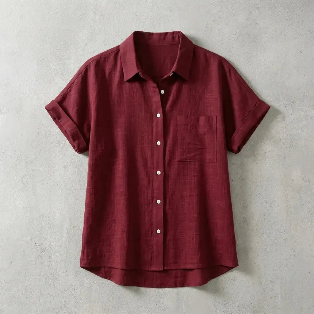 Folded burgundy red linen short-sleeve shirt with chest pocket and mother-of-pearl buttons, laid flat on concrete background