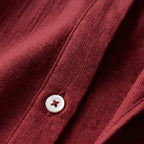 Close-up of woven linen texture on a burgundy shirt showing a mother-of-pearl button and fine stitching detail