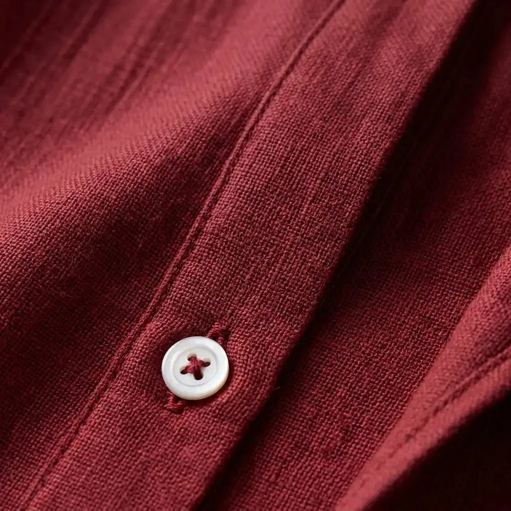 Close-up of woven linen texture on a burgundy shirt showing a mother-of-pearl button and fine stitching detail