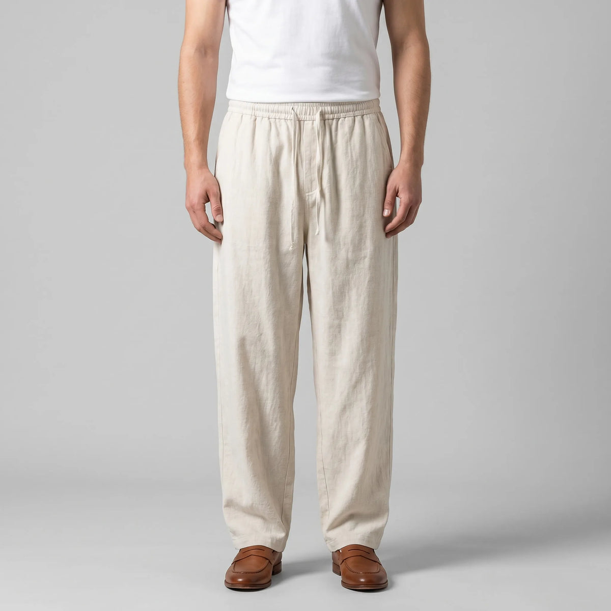 Front view of man wearing beige baggy linen pants with drawstring waist, styled with a white singlet and tan leather loafers