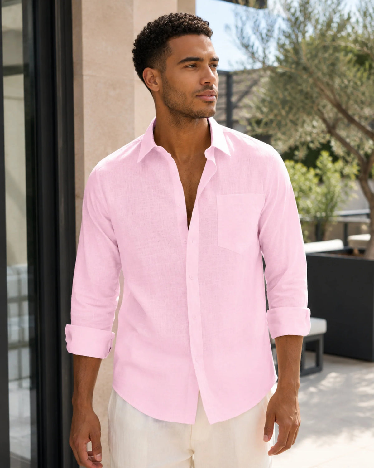 Man in open-collar pink linen shirt with sleeves rolled, worn over cream pants outside a modern home