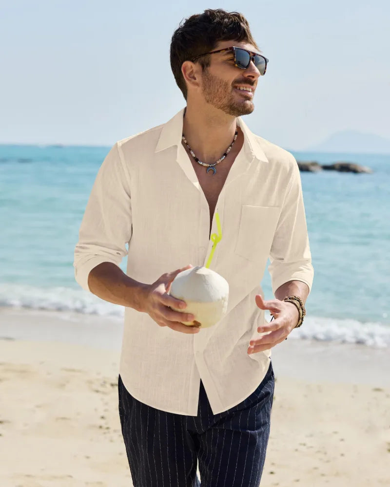 Man at the beach in an off-white linen shirt, collar open, sleeves rolled, holding a coconut drink