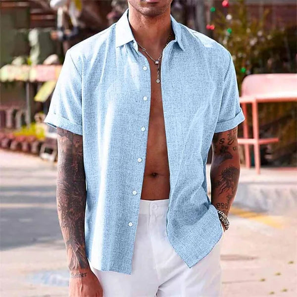 Man wearing a light blue cotton linen short-sleeve shirt, unbuttoned, paired with white trousers on a city street
