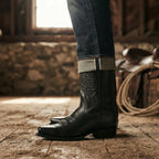 Model wearing black leather cowboy boots with cuffed denim jeans, standing on a wooden floor in a barn setting