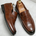 Close-up pair of brown leather penny loafers with shoe trees, showing stitched apron toe and stacked heel