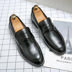 Black leather penny loafers with shoe trees on a chequered surface, showing glossy finish and apron stitching
