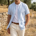 Man wearing a pale blue linen short sleeve shirt unbuttoned over a white tee and cream pants outdoors