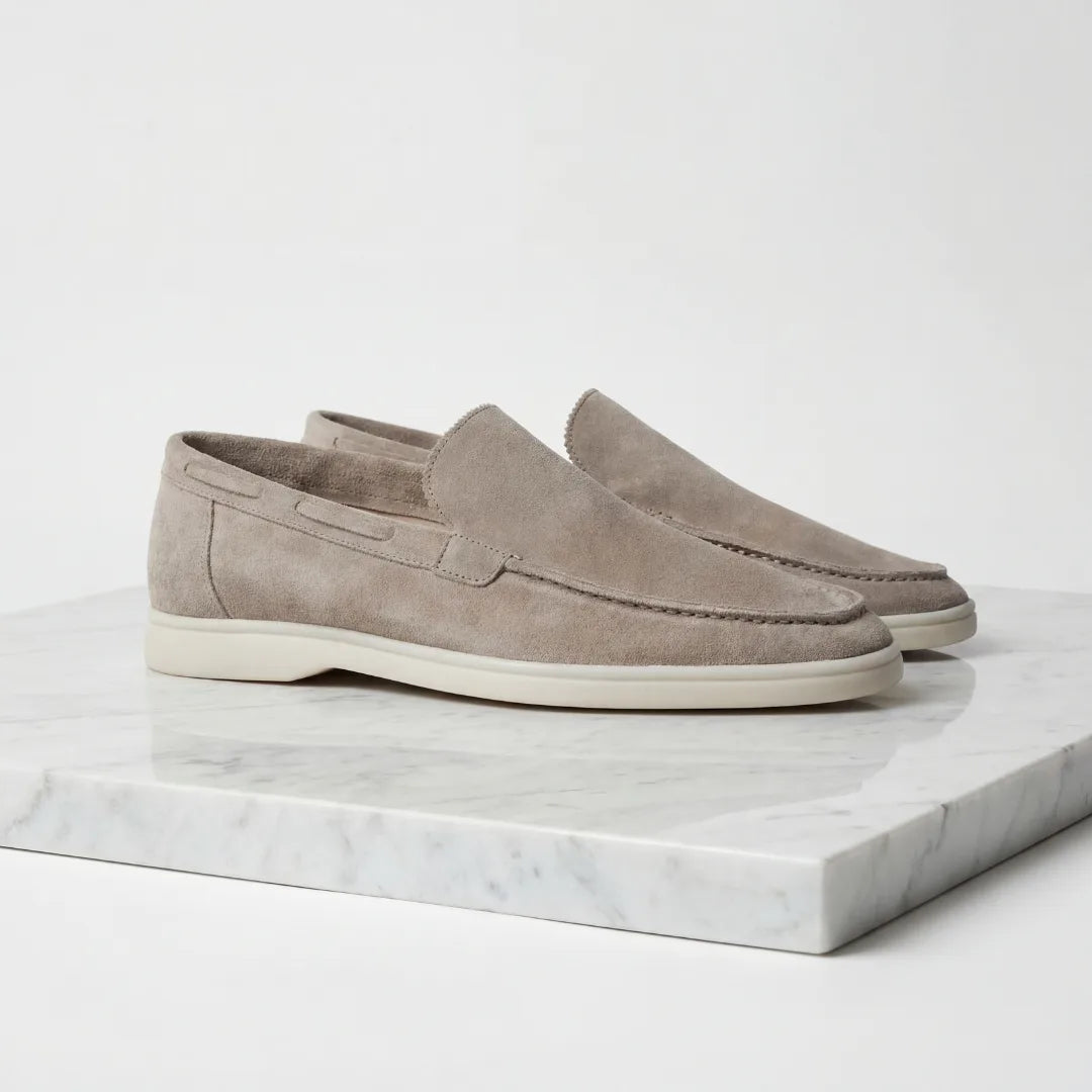 Side profile of a pair of light taupe suede loafers with white rubber soles on a marble plinth