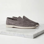 Pair of taupe suede loafers shown slightly angled on a white marble surface