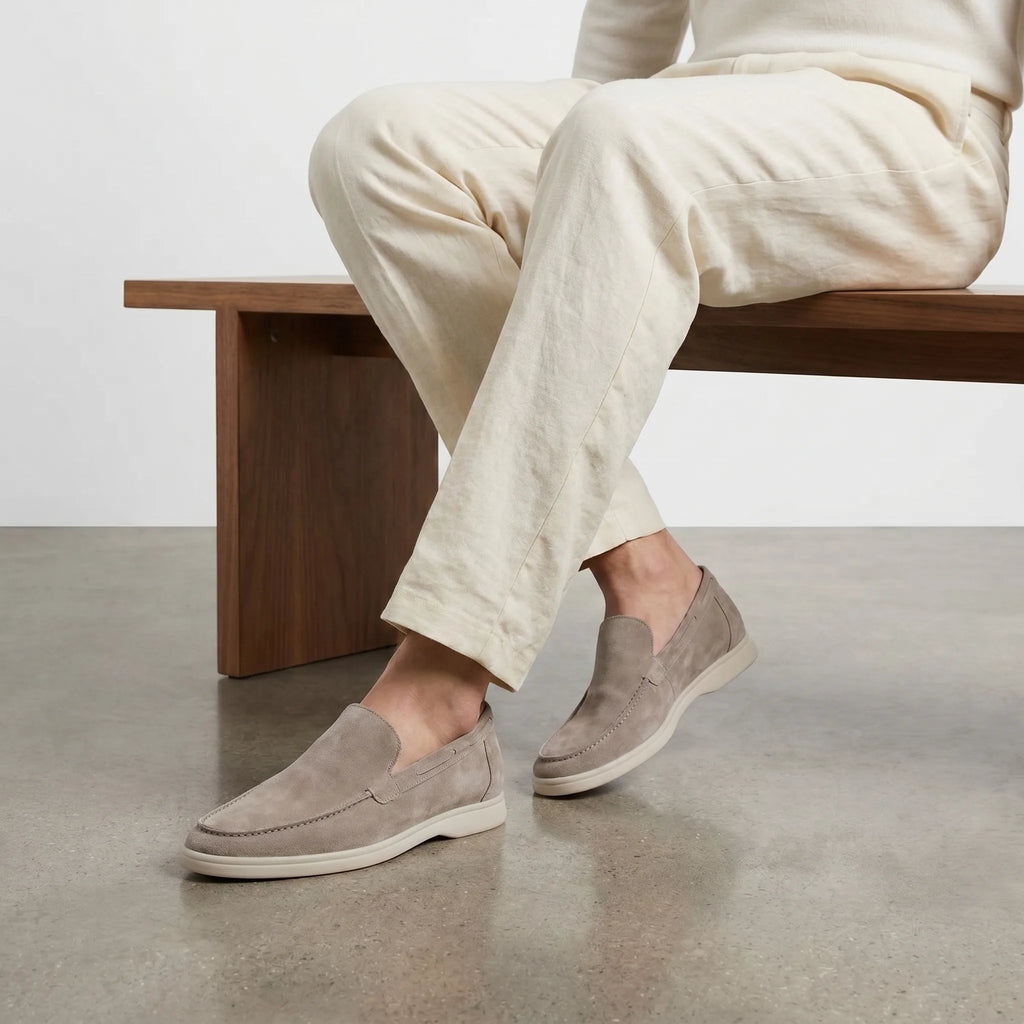 Model wearing taupe suede loafers with cream trousers while seated on a wooden bench