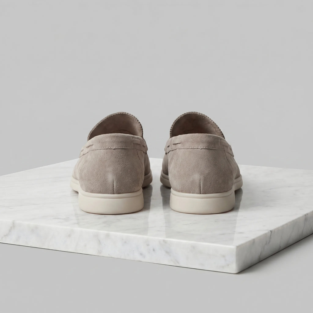 Rear view of a pair of taupe suede loafers showing the heel seam and white outsole on marble