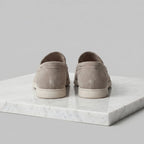 Rear view of a pair of taupe suede loafers showing the heel seam and white outsole on marble