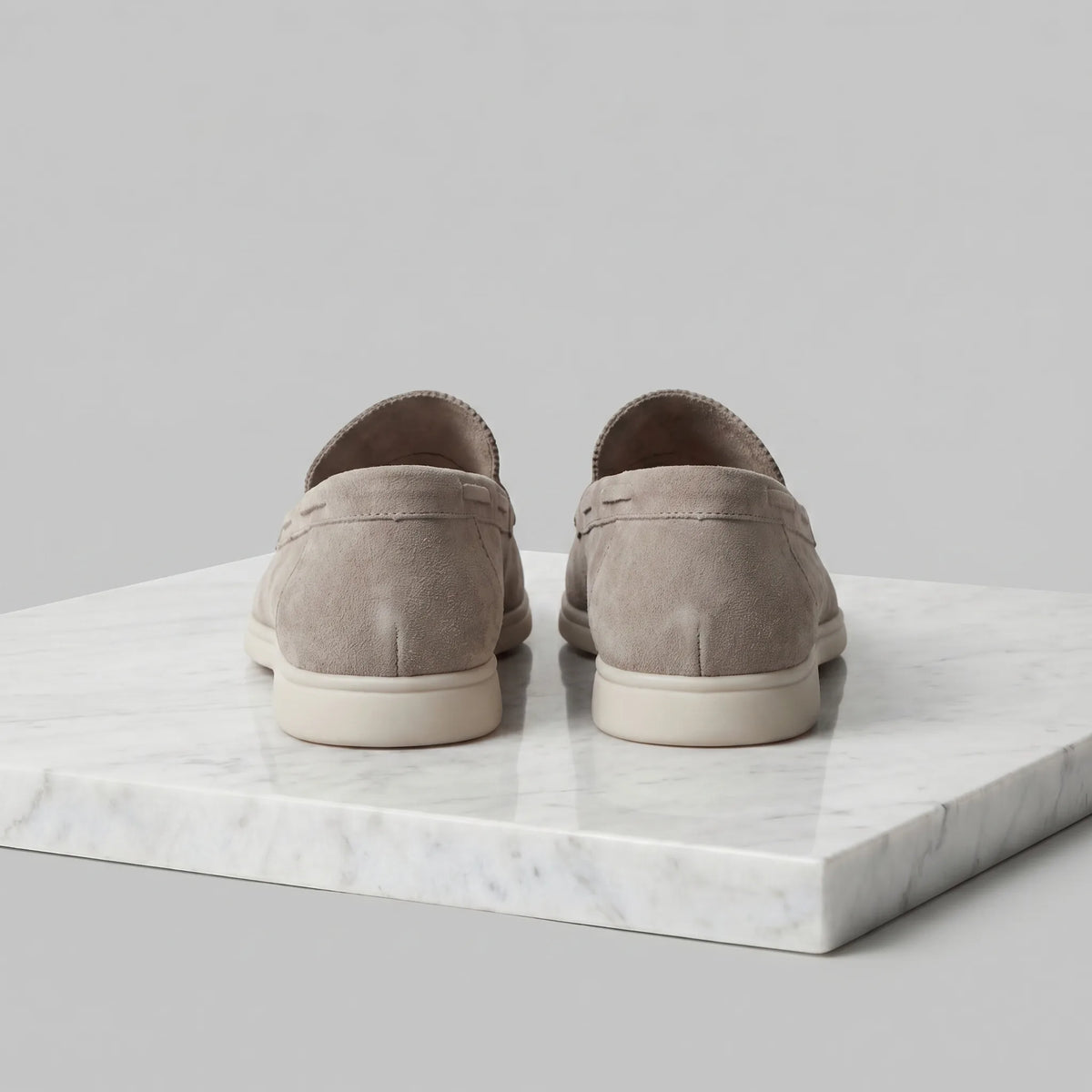 Rear view of a pair of taupe suede loafers showing the heel seam and white outsole on marble