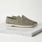 Side profile of a pair of olive green suede loafers with white soles on a marble plinth