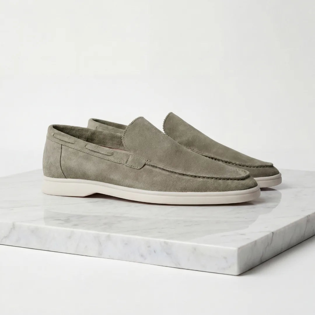 Side profile of a pair of olive green suede loafers with white soles on a marble plinth