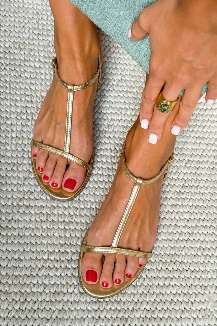 Overhead view of metallic gold T-strap flat sandal with buckle ankle strap, worn with red pedicure on a woven rug