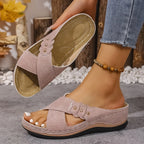 Overhead shot of navy blue wedge sandals with gold stud trim resting beside a bare foot near pebbles