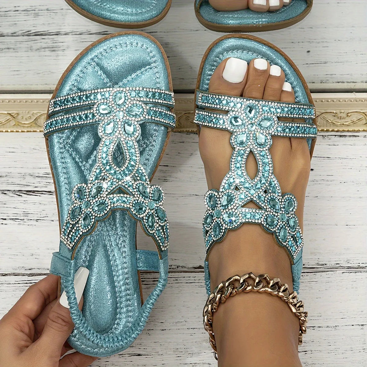 Overhead view of pale blue jewelled sandals with rhinestone floral straps, one held by hand to show the quilted footbed