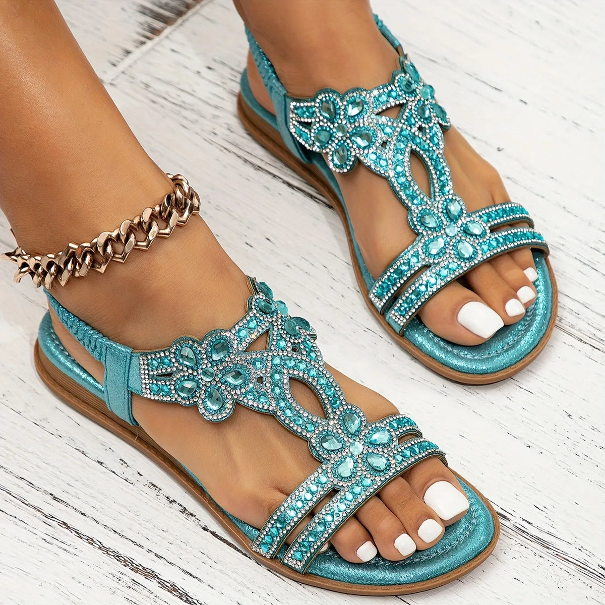 Side angle of navy blue jewelled sandals worn outdoors on a wooden deck, with gold chain anklet detail