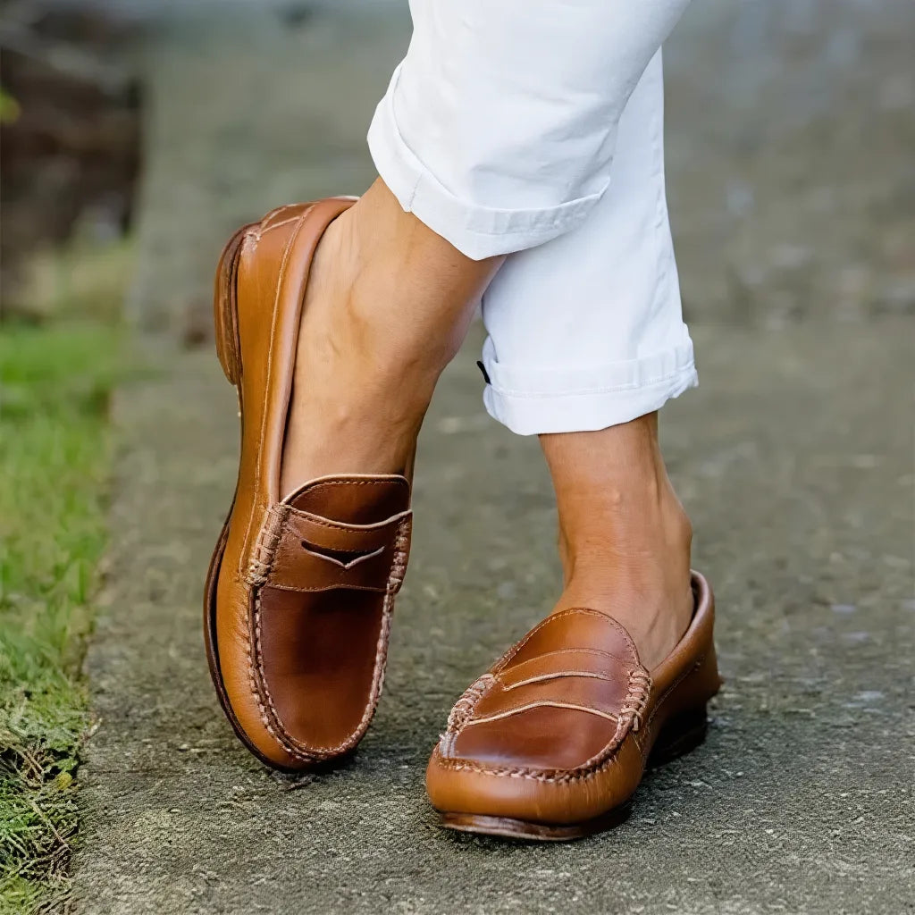 Close-up of tan leather penny loafers worn with white cuffed trousers, showing glossy finish and stitched apron toe.