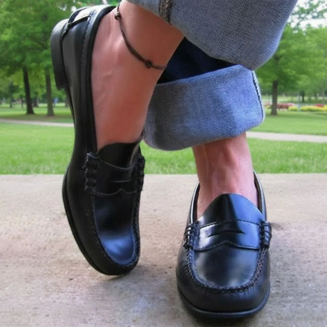 Black leather penny loafers worn outdoors with grey cuffed trousers, showing the classic saddle strap and low heel.
