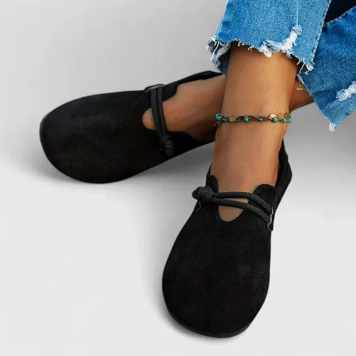 Side profile of black suede flats worn on bare feet, showing the rope strap knot and low cut vamp