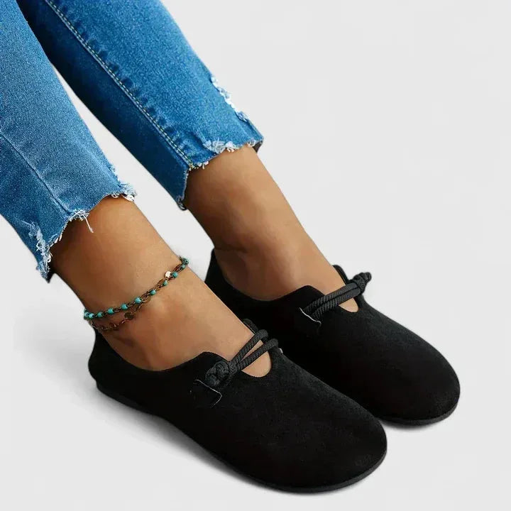 Black suede flats worn on crossed ankles with ripped denim, showing the rope strap and sleek silhouette