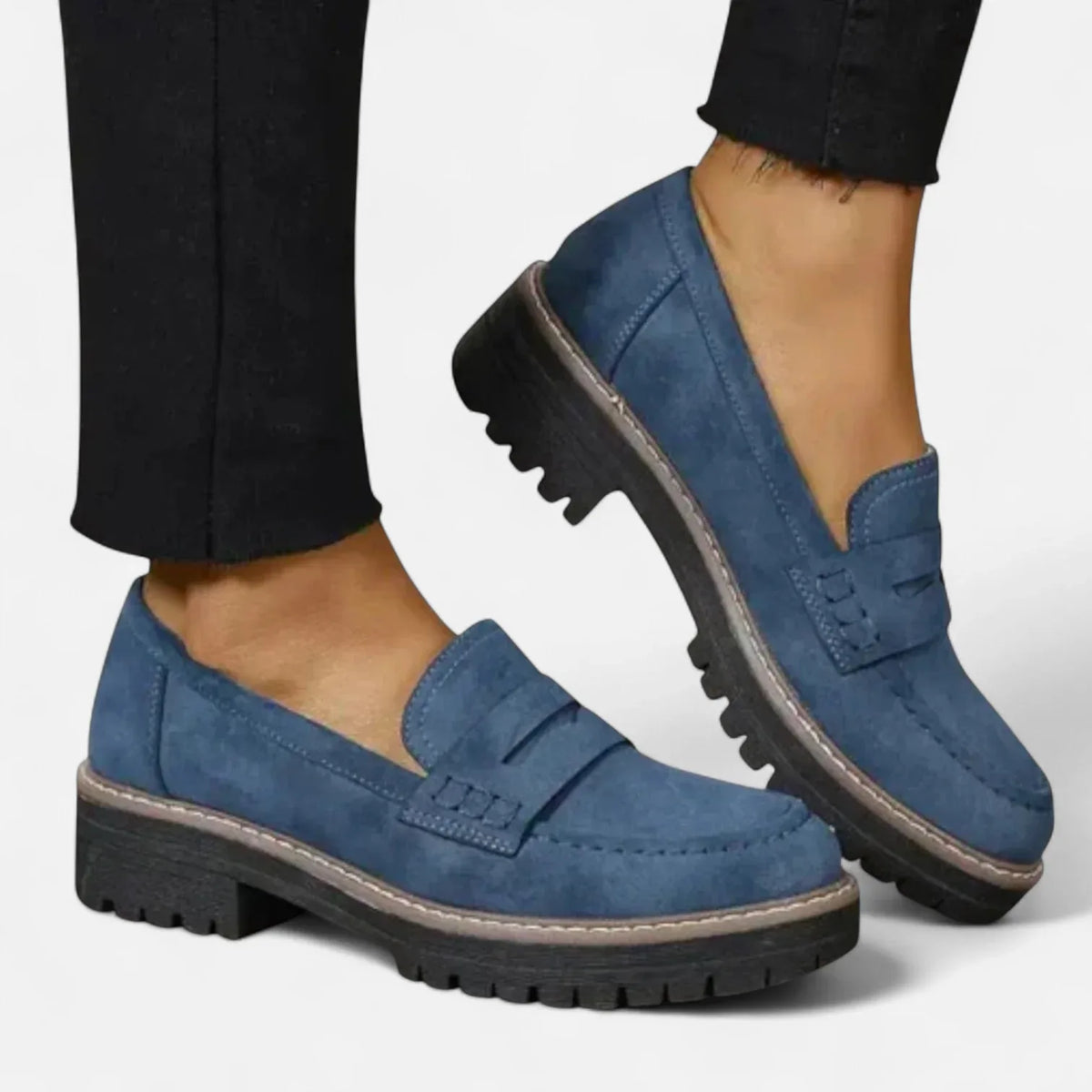 Side view of a navy blue suede penny loafer with chunky black lug sole, worn with cropped black trousers