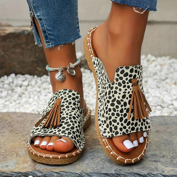 Pair of dalmatian-print sandals with suede tassel accent and stitched tan sole shown on stone flooring