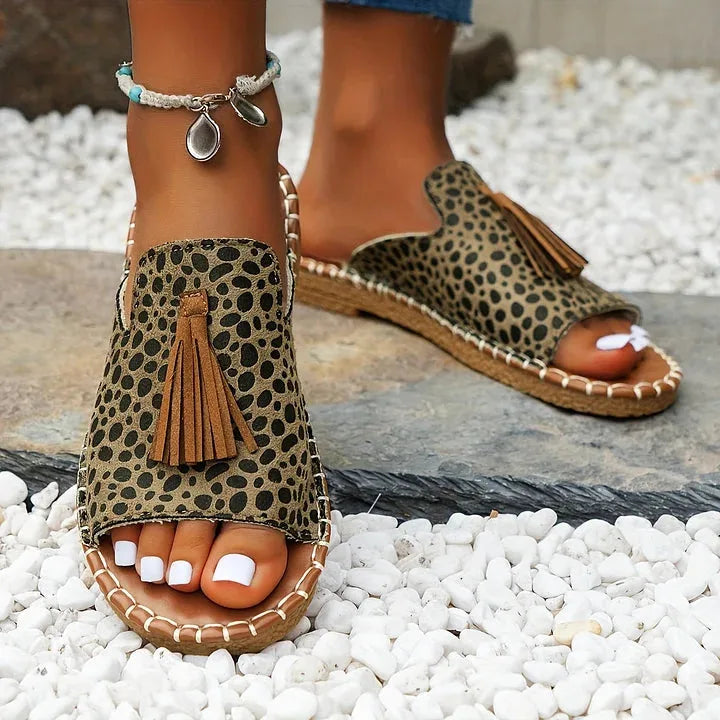 Close-up front view of leopard-print sandals with tan tassel and woven jute-style sole on pebbled ground